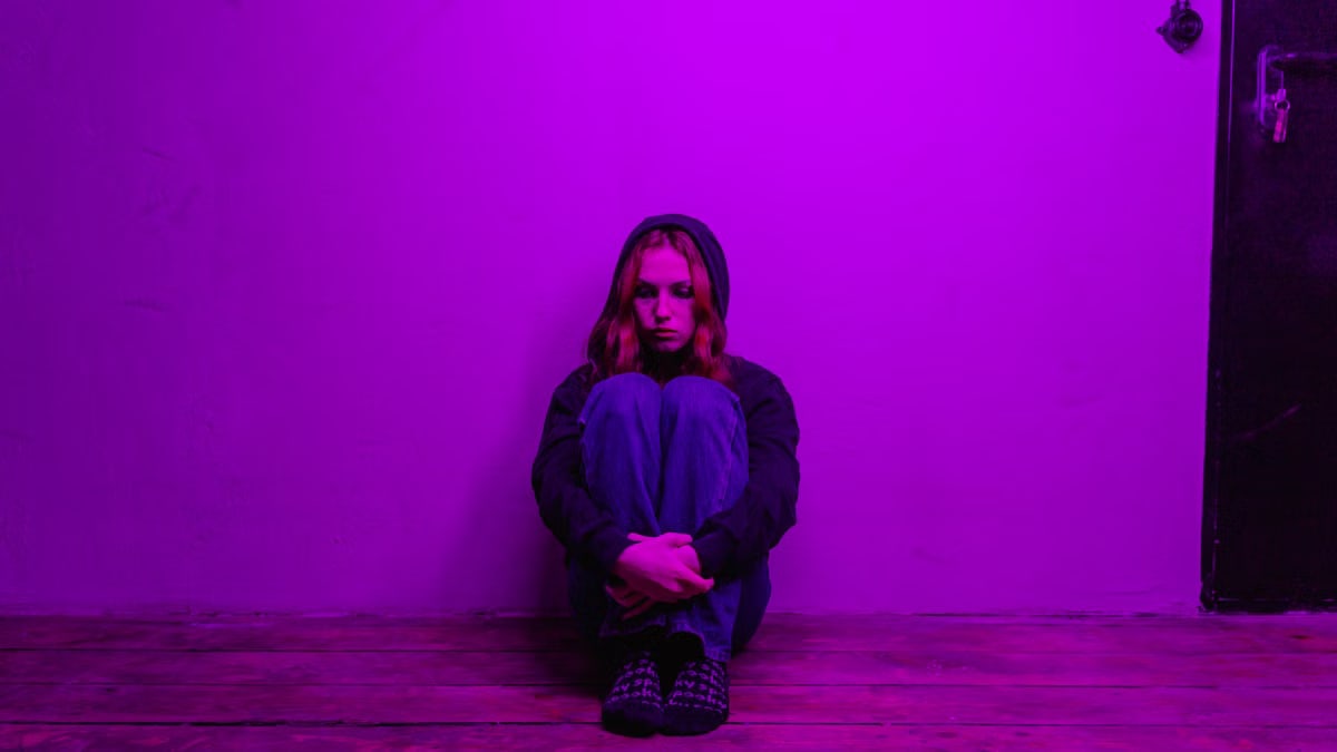 A woman in a hoodie sits alone on the floor, illuminated by purple light indoors.