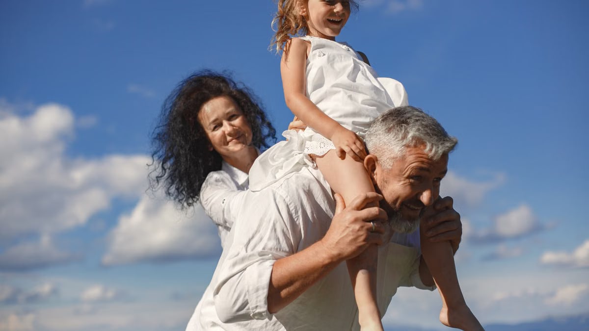 Grandparents playing with granddaughter outdoors under bright blue sky.