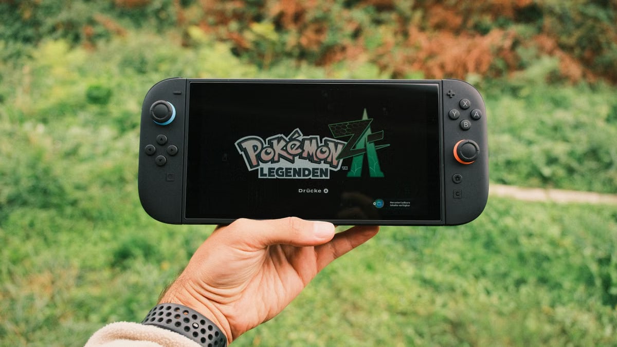 A hand holding a handheld gaming console displaying the Pokémon Legends game screen outdoors.
